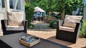 An outdoor seating area with wicker chairs and a table, a small garden, and a building with a sign in the background. - Sport Hotel Kenzingen (Kenzingen)