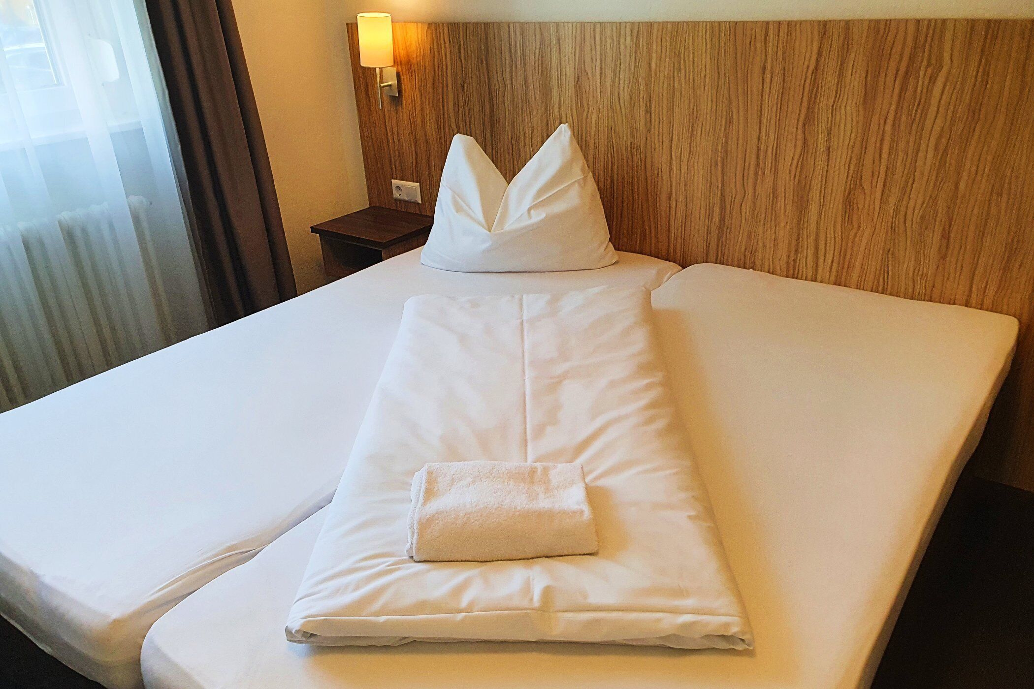 A neatly made bed with white linens, a folded towel, and a decorative pillow on a wooden headboard.