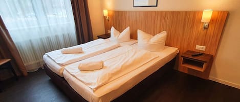 Uma cama de casal com roupa de cama e almofadas brancas, uma cabeceira de madeira, duas mesas de cabeceira com abajures e uma janela com cortinas.