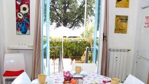 Uma área de refeições com uma mesa posta para dois, com toalha de mesa florida, pratos, xícaras e um jarro de suco.