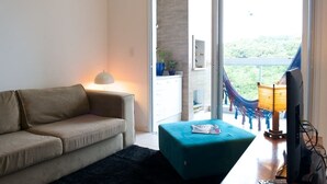 A modern living room with a sofa, a small table, and a television. There is a balcony with a hammock and a view of greenery. - Sea Front, Front Native Forest (Florianópolis)