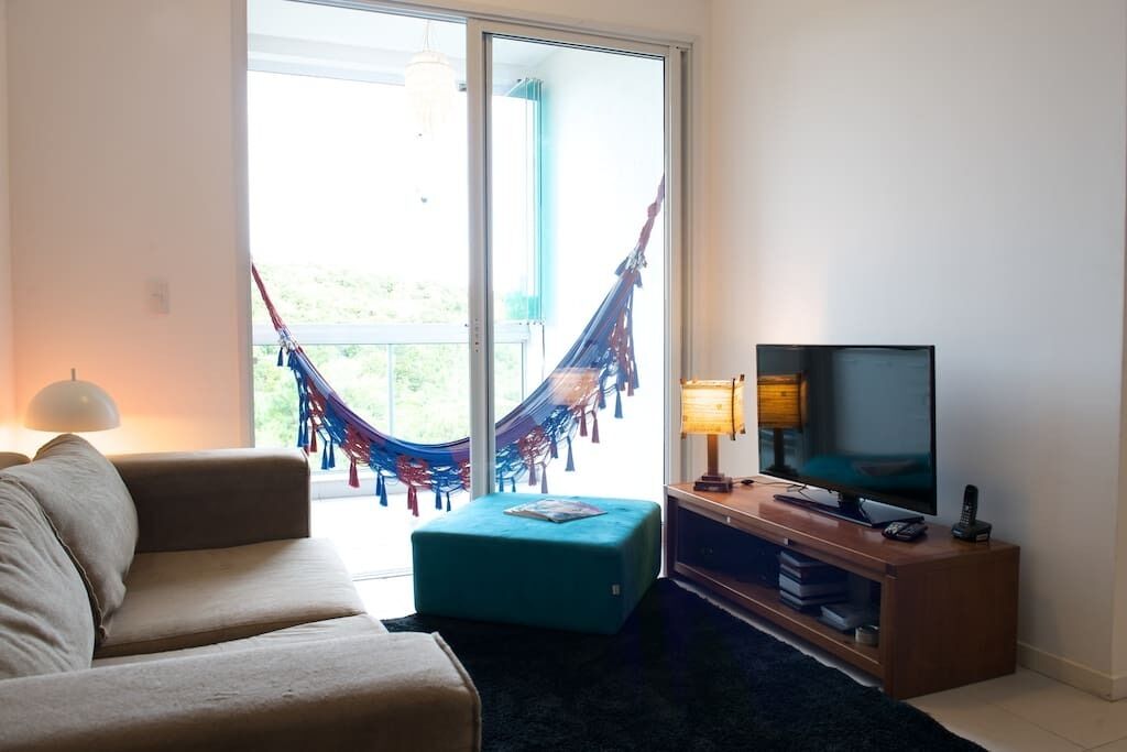 Una sala de estar con un sofá, un televisor, un pequeño reposapiés y una hamaca junto a una puerta corrediza de vidrio.