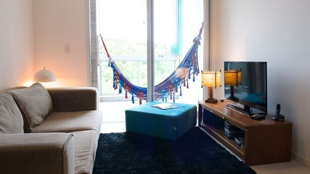 A living room with a sofa, a TV on a wooden stand, a hammock, and a balcony with a view. - Sea Front, Front Native Forest (Florianópolis)