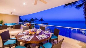 Een eetruimte met een ronde tafel voor vier personen, omringd door rieten stoelen met blauwe kussens, met uitzicht op een zwembad en de oceaan tijdens de zonsondergang.