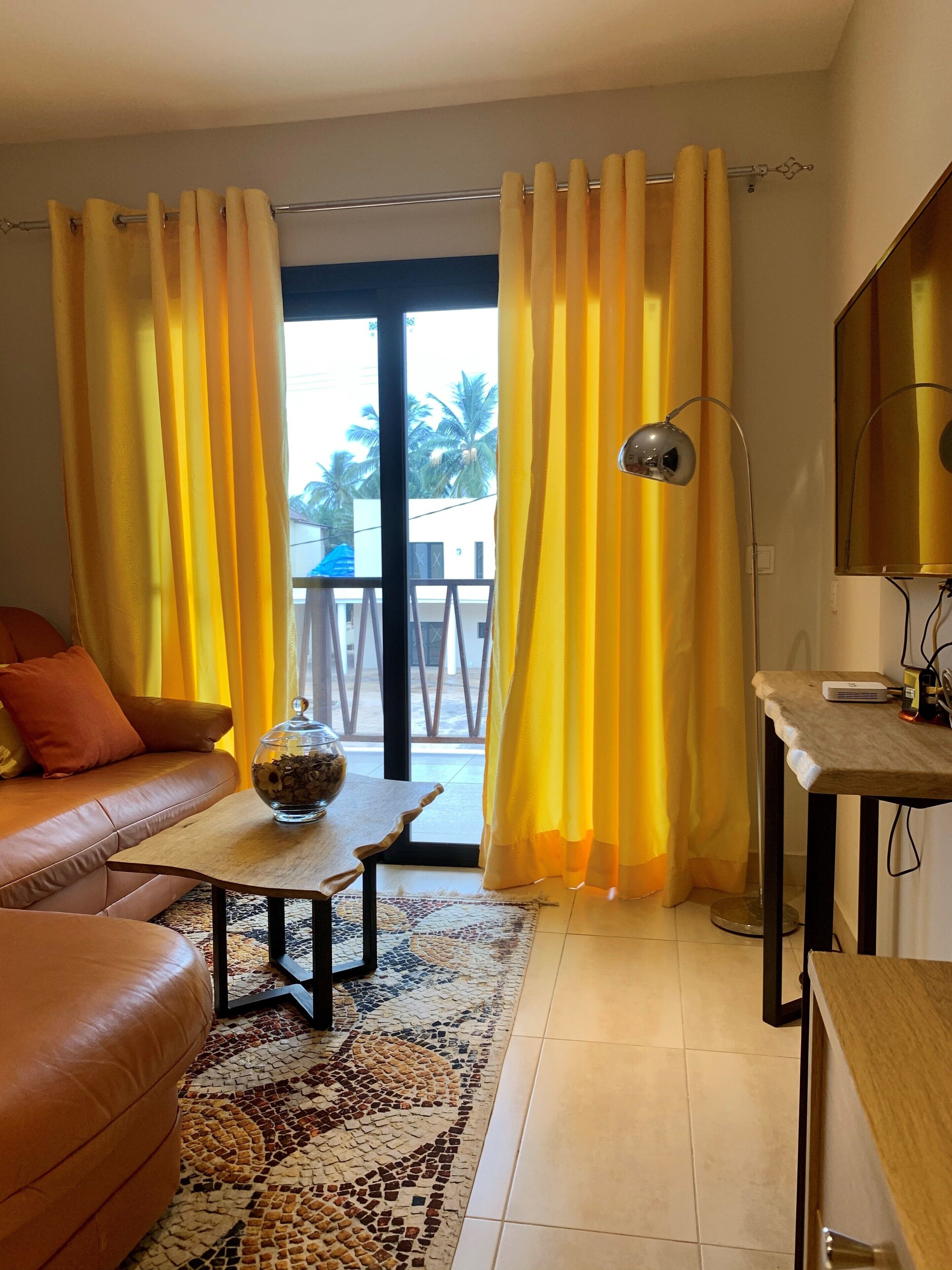 A living room with a sofa, a coffee table with a decorative bowl, yellow curtains, a floor lamp, and a view of palm trees outside.