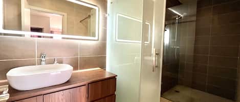 Un baño moderno con un espejo grande, un lavamanos blanco, armarios de madera y una ducha con mampara de vidrio.