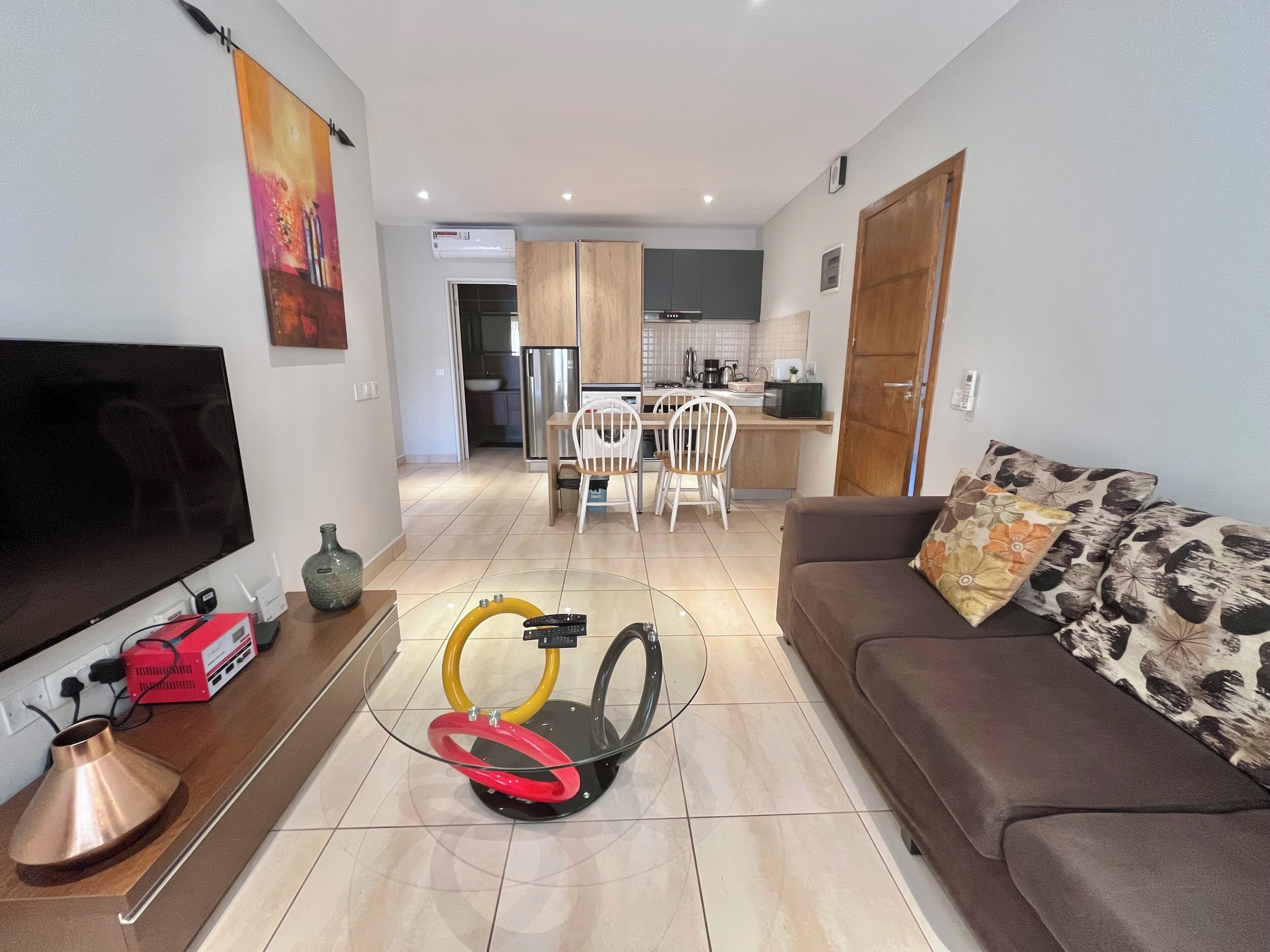 A living room with a TV, a glass coffee table, a sofa, and a kitchen in the background.
