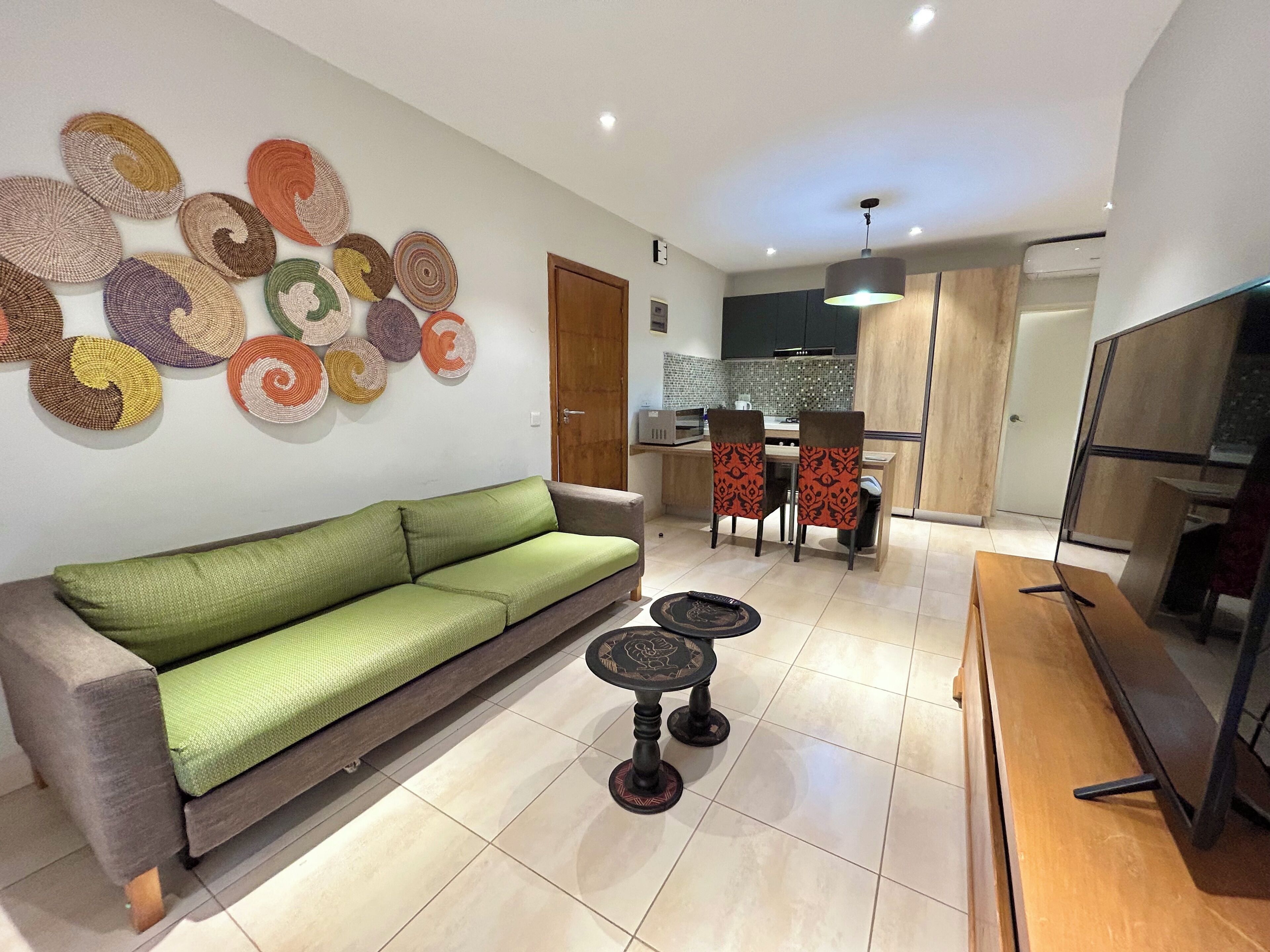 A modern living room with a green sofa, a dining area with patterned chairs, and a kitchen in the background.