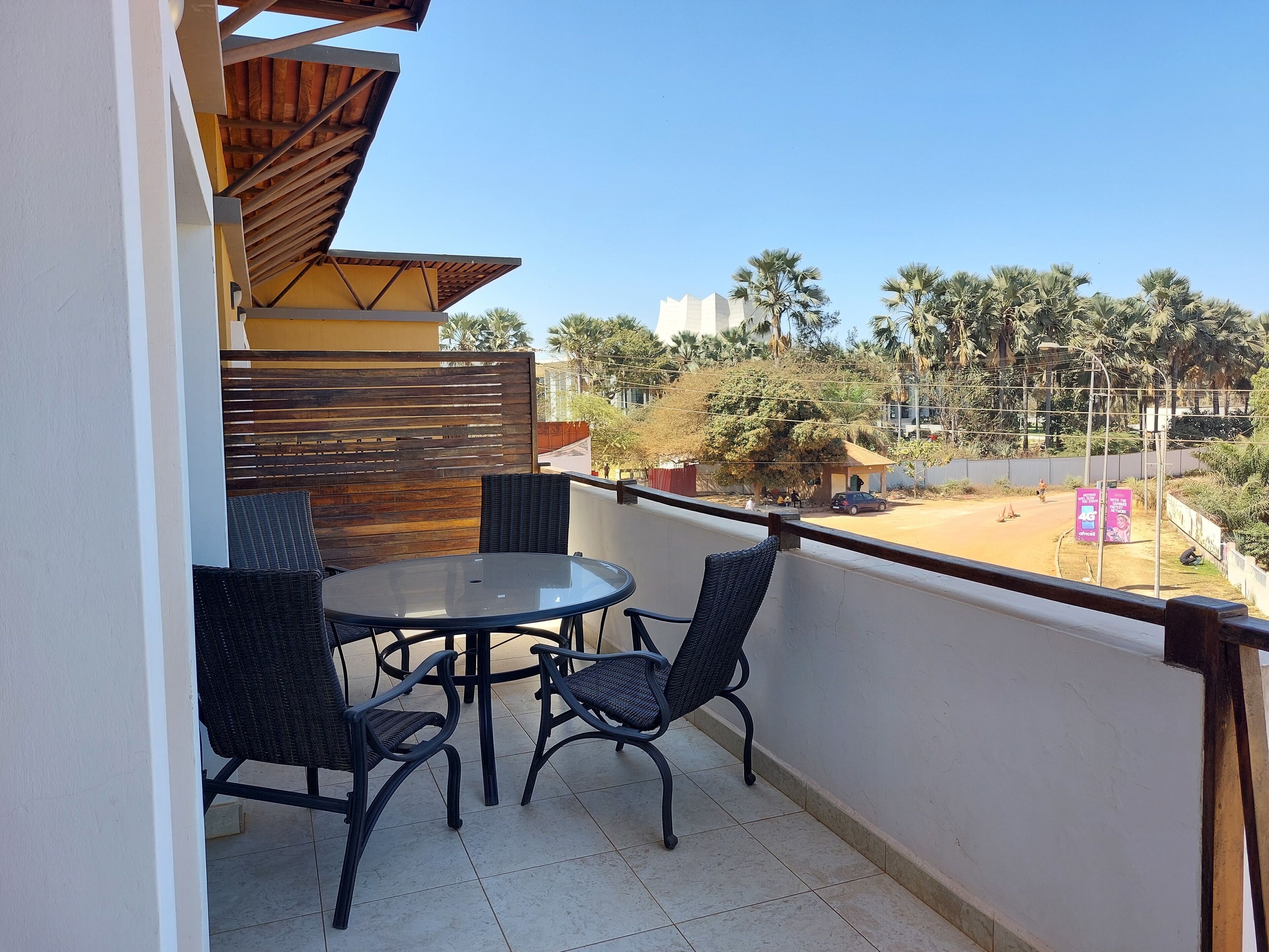 A balcony with a round table and four chairs, a wooden slatted partition, and a view of palm trees and buildings in the distance.