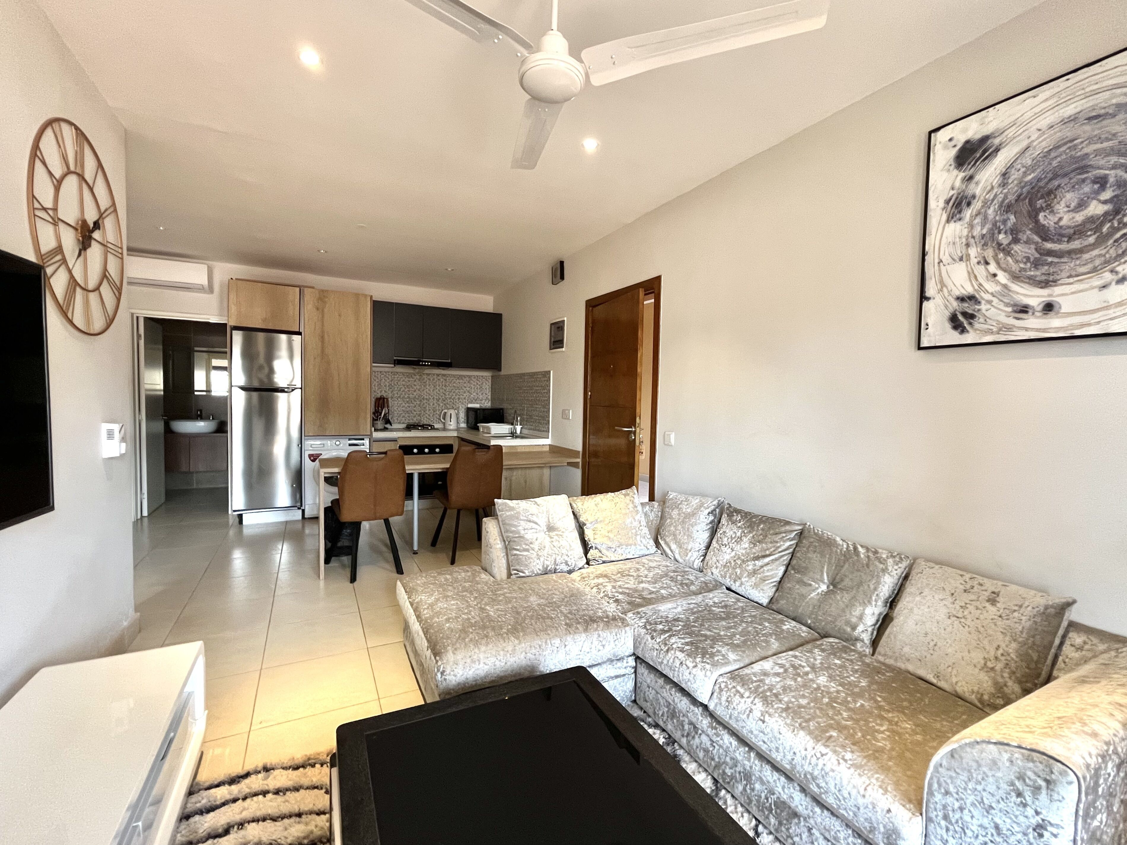 A modern living room with a sectional sofa, a coffee table, and a TV. There is a kitchen in the background with a refrigerator, microwave, and cabinets.
