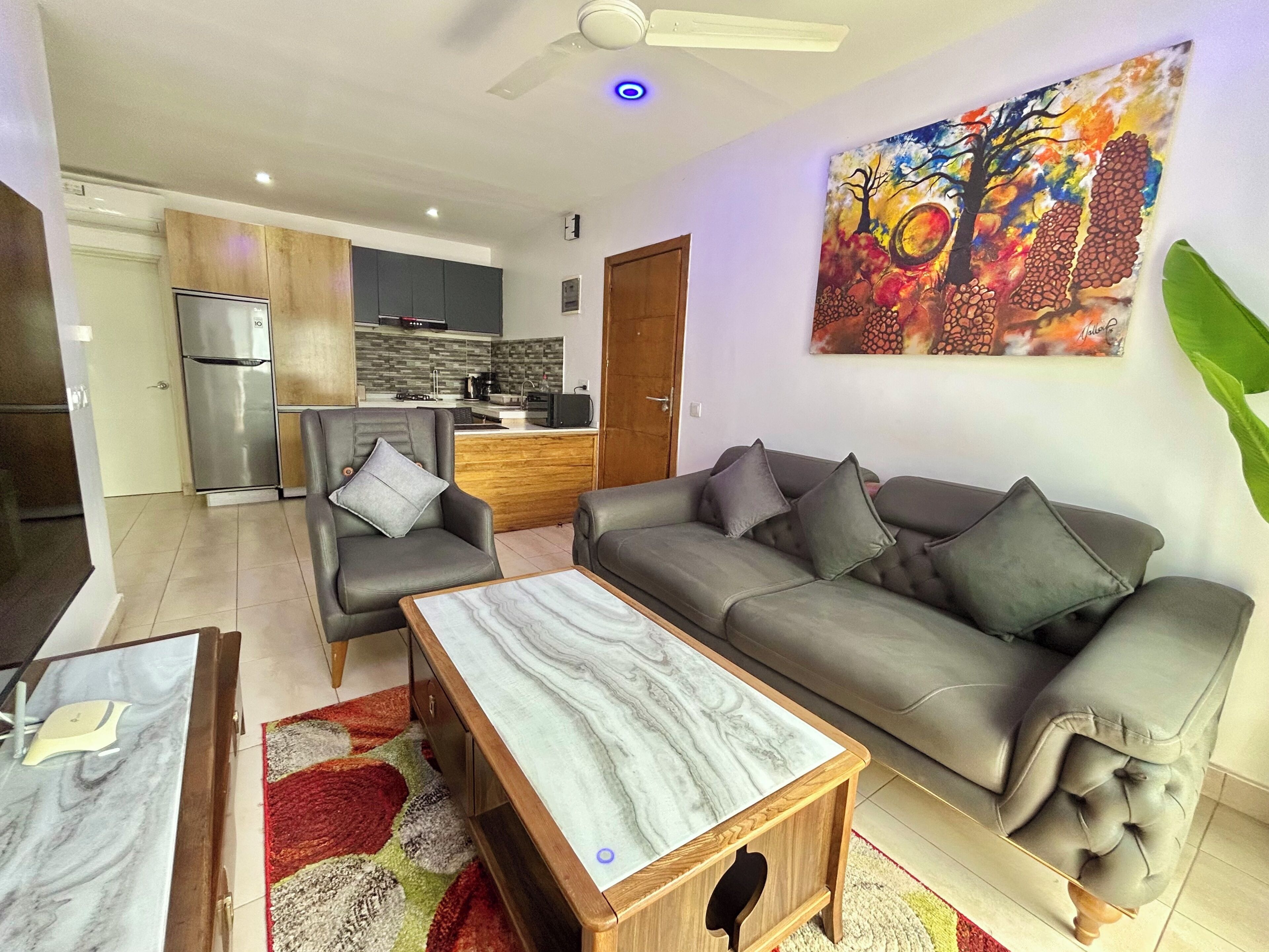Una sala de estar moderna con sofá, sillón y mesa de centro. Se ve una cocina con refrigerador y microondas al fondo.