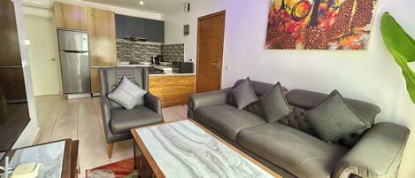 Una sala de estar moderna con sofá, sillón y mesa de centro. Se ve una cocina con refrigerador y microondas al fondo.