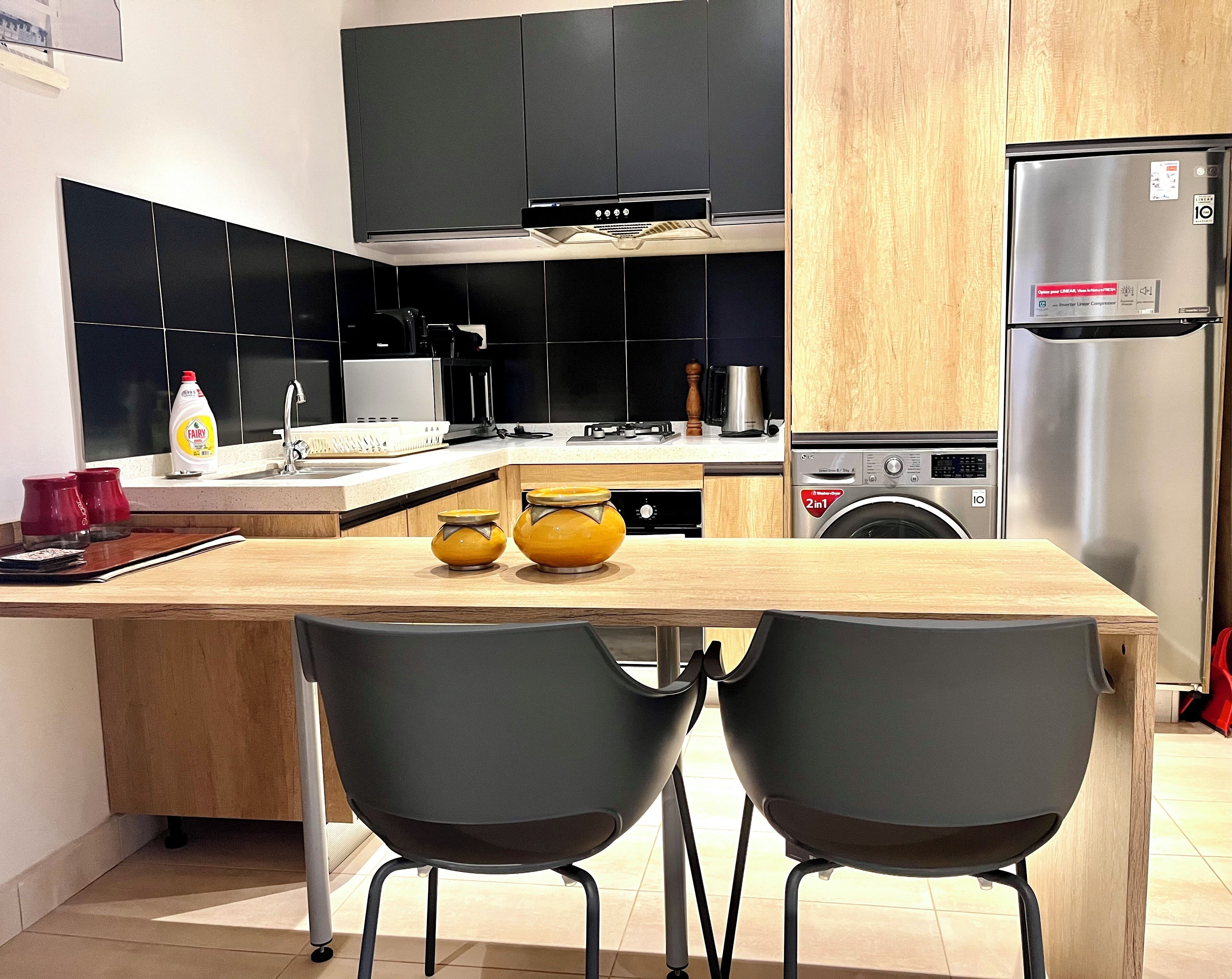 Een moderne keuken met zwarte kasten, een roestvrijstalen koelkast, een wasmachine en twee zwarte stoelen aan een houten tafel.