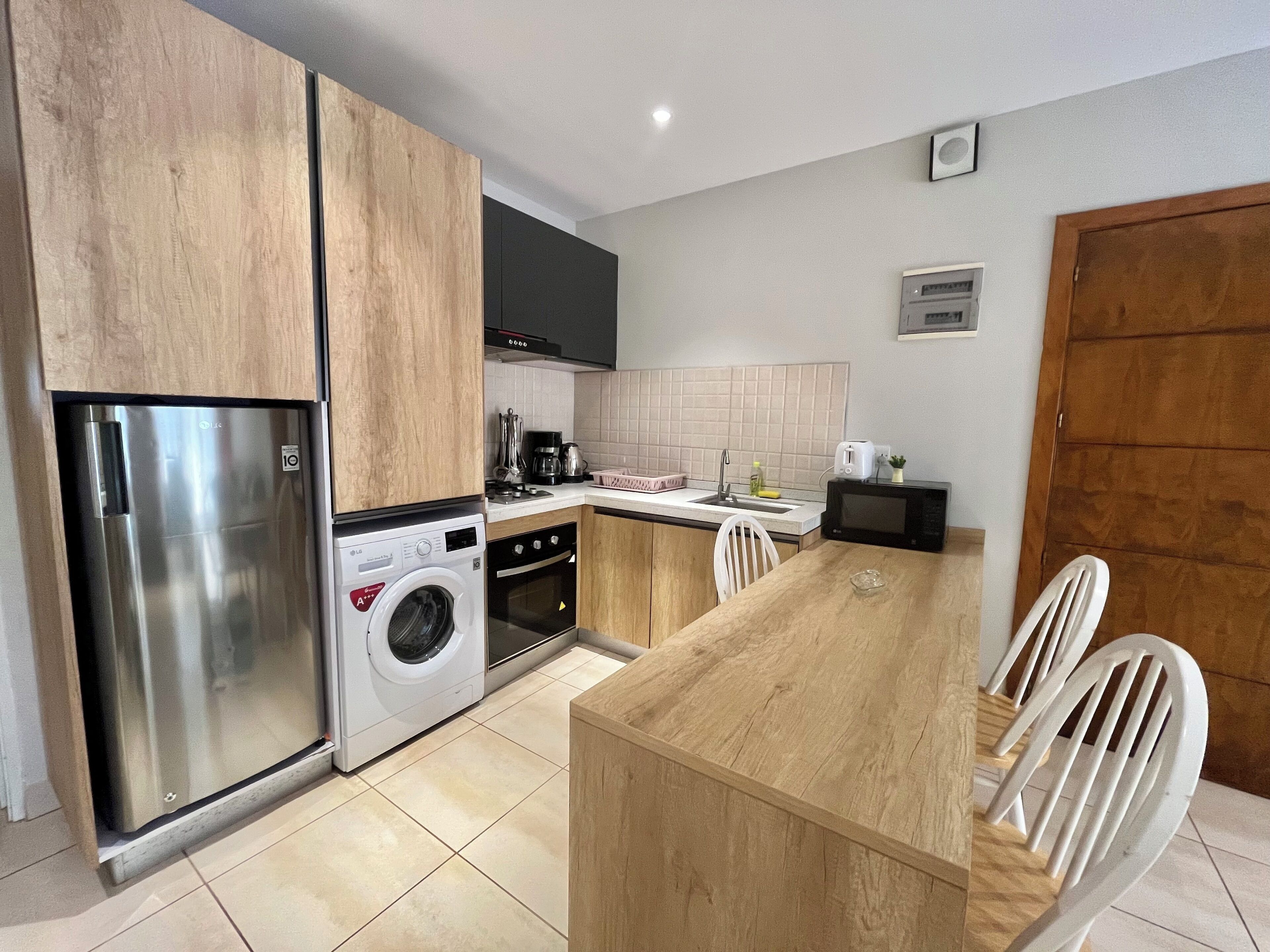 A compact kitchen with a stainless steel refrigerator, a washing machine, an oven, a microwave, and a sink. There is a wooden dining table with four chairs.
