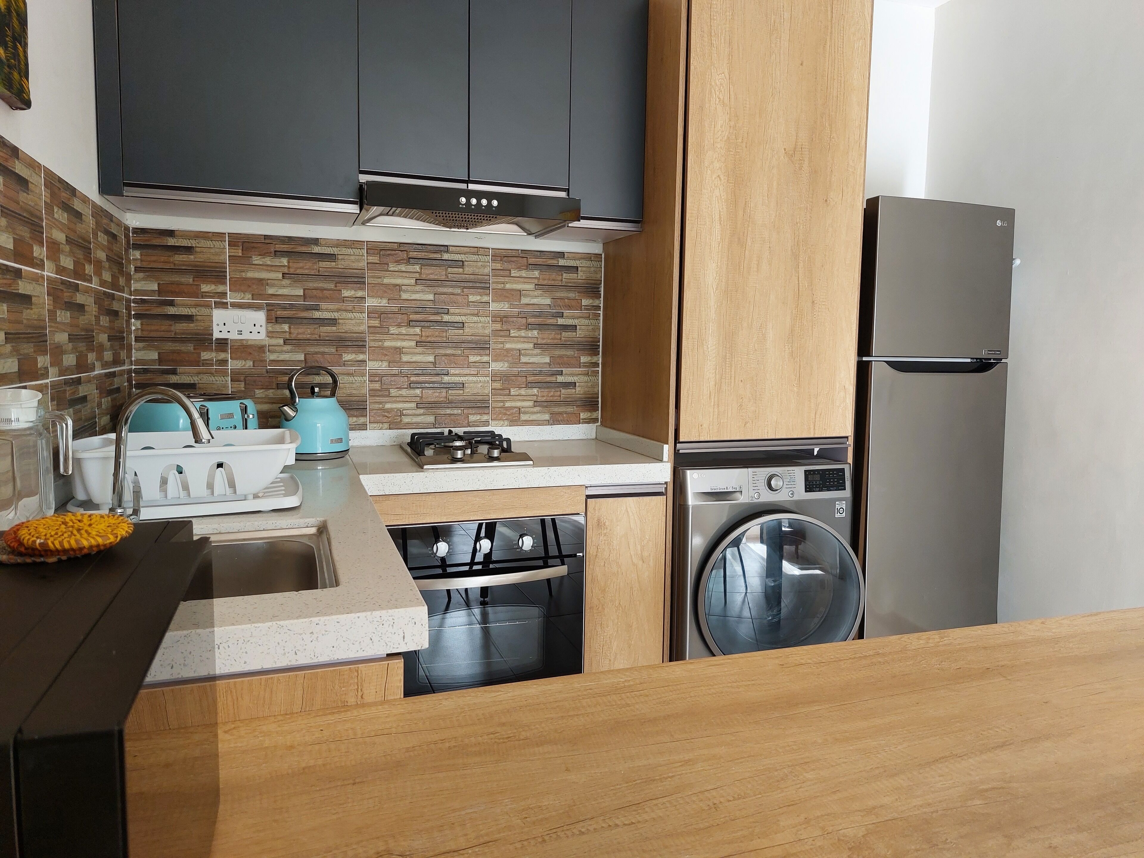A modern kitchen with grey cabinets, a stainless steel washing machine, a blue kettle, and a gas stove.