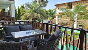 A balcony with wicker furniture, a glass table, and a view of a swimming pool and greenery. - Forest View Apartments (Serrekunda)