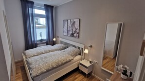 A modern bedroom with two beds, a bedside table with a lamp, a small bedside table with a plant, a large mirror, and a window with curtains. - Vacation apartment Ahr-Flair (Bad Neuenahr-Ahrweiler)