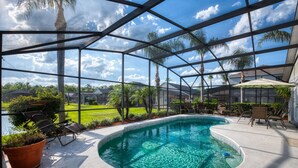 A swimming pool with a glass enclosure, surrounded by lounge chairs and umbrellas, with a view of a residential area and palm trees. - NEW LISTING Stunning Villa - Amazing Value - Lake Berkley (Kissimmee)