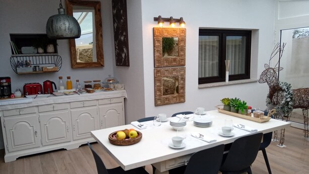 Ein Essbereich mit einem weißen Tisch, gedeckt für vier Personen, umgeben von schwarzen Stühlen. Im Hintergrund ist eine Küchenzeile mit einem weißen Schrank und einer Pendelleuchte zu sehen.
