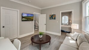 A spacious living room with a round coffee table, a sofa, and a TV mounted on the wall. There is a staircase leading to another level, and a door leading to a bedroom.