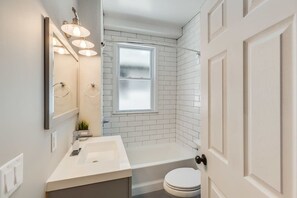 A bathroom with a white sink, mirror, and toilet, a bathtub, and a window.