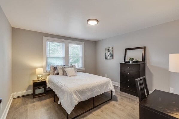 A bedroom with a bed, bedside table, dresser, and a window with blinds.