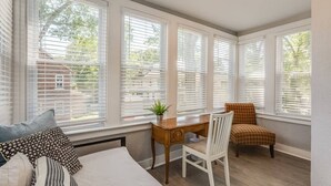A room with a bed, a desk, and a chair near large windows with blinds.