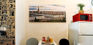 A compact kitchen with a microwave on top of a fridge, a table with a striped tablecloth, and a cityscape painting on the wall.