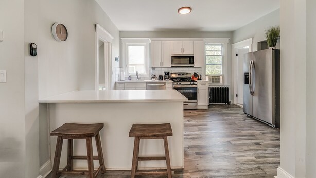 A modern kitchen with white cabinetry, a central island with stools, stainless steel appliances, and a view of the adjacent living area. - 4BR/2BA Duplex Apt in Downtown Montclair w/Parking (Montclair)