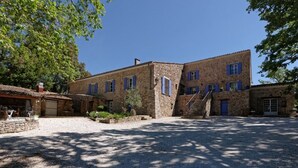Un bâtiment en pierre avec des volets bleus, une cour en gravier et un escalier menant à l’entrée.