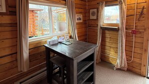 Puinen mökki, jonka sisustuksessa on pöytä, kaksi verhoilla varustettua ikkunaa, seinään kiinnitetty valaisin ja kehystetyt valokuvat.