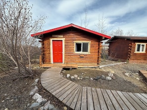 A wooden cabin with a red door and a window, surrounded by a wooden deck and natural landscape.