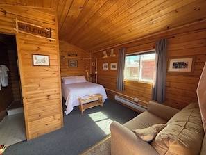 Een houten kamer met een bed, een houten bankje, een bank en een raam, plus een welkomstbord.