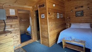 A wooden cabin room with a bed, a small table, a sofa, and a door leading to another room. - Earthsong Lodge -Two Room Cabin (Healy)