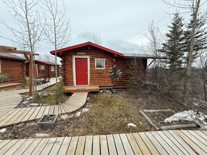 A log cabin with a red door, a small window, and a metal windmill in the yard.