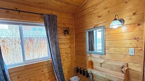 L’intérieur d’un chalet en bois, avec un banc, une fenêtre avec des rideaux, un miroir fixé au mur, un luminaire fixé au mur et une petite poubelle.