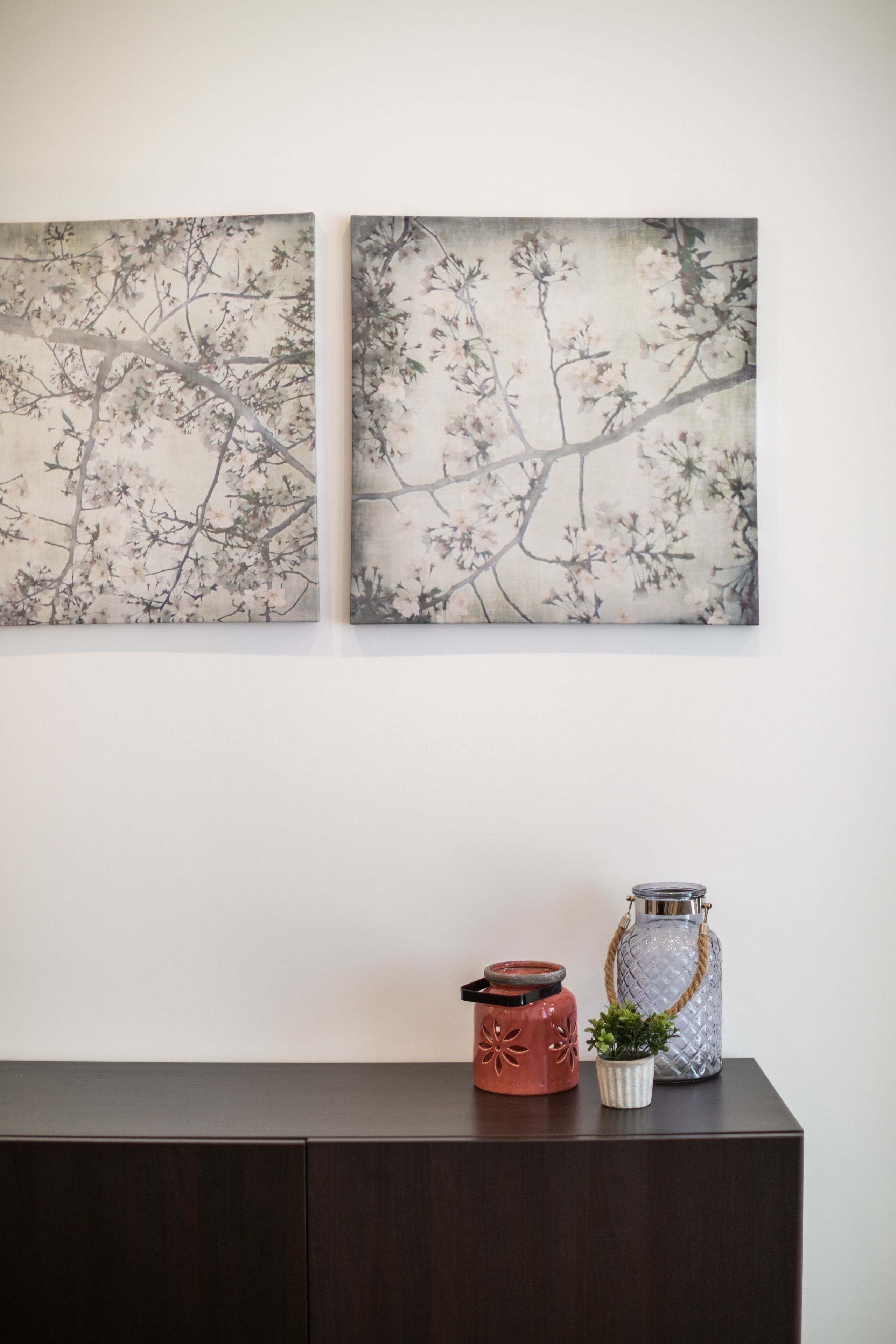Dos cuadros colgados en la pared con ramas y flores, un mueble de madera oscura y una mesita con una maceta roja y un tarro de vidrio con asa de cuerda.