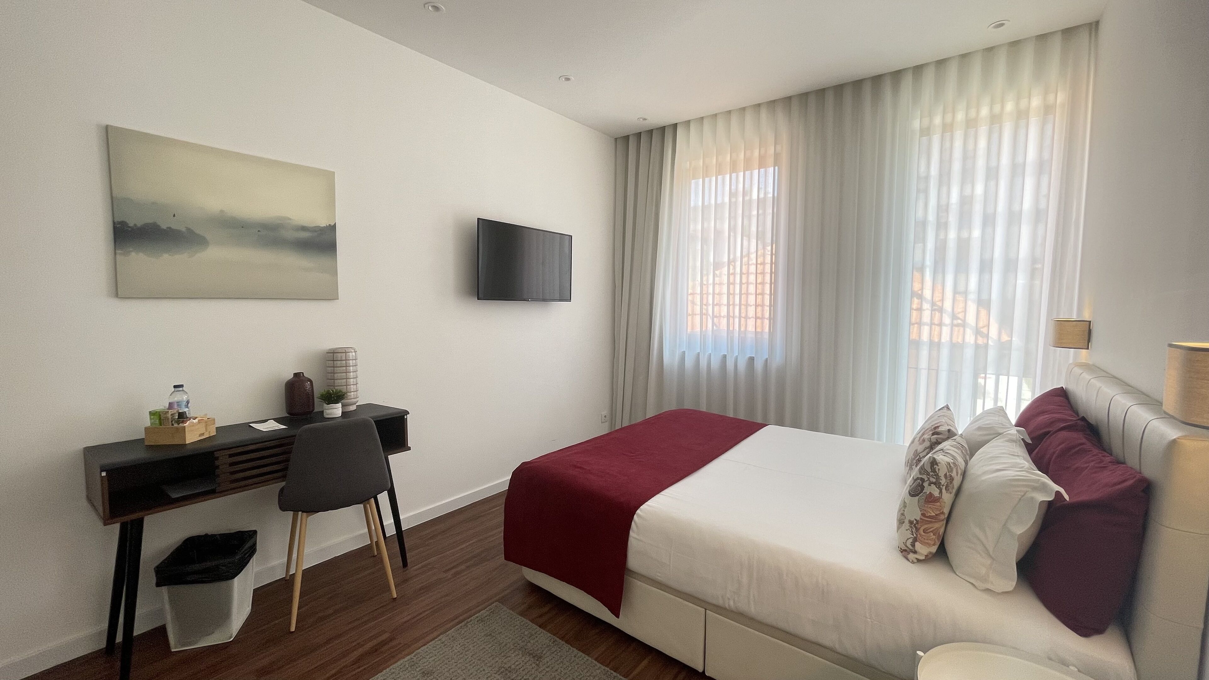 Quarto de hotel com cama, escrivaninha, cadeira, televisão e janela com cortinas.