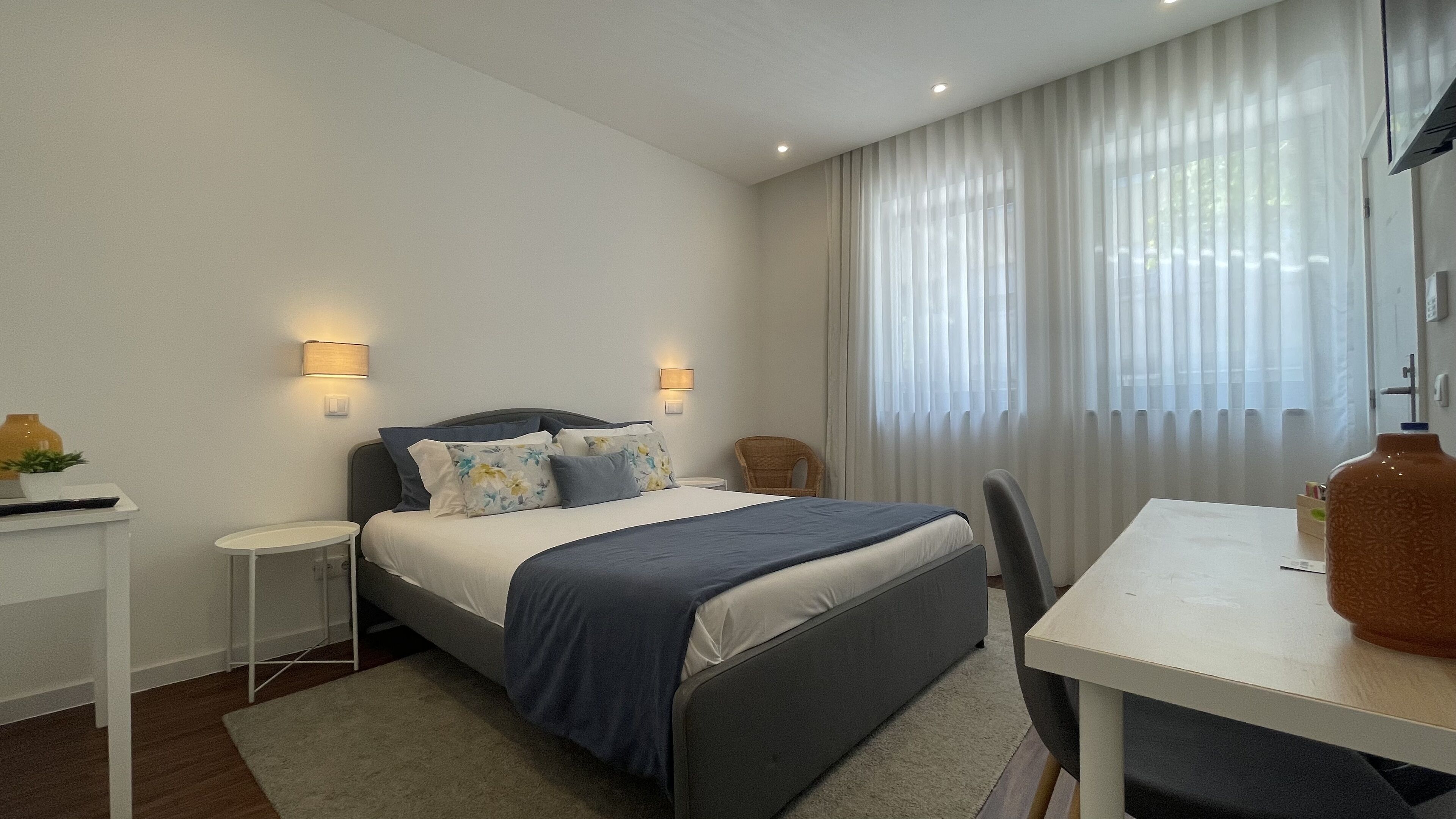 A modern hotel room with a neatly made bed, a small side table, a chair, and a desk with a vase.