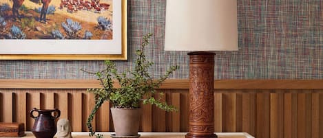 A wooden console table with a woven design, a potted plant, a ceramic pitcher, and a framed painting of a cowboy on a horse.