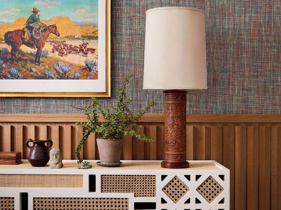 A wooden console table with a woven design, a potted plant, a ceramic pitcher, and a framed painting of a cowboy on a horse.