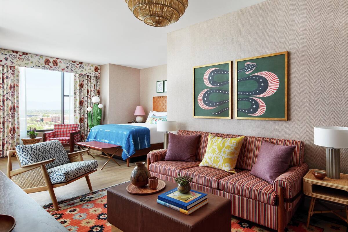 A hotel room with a bed, a striped sofa, a patterned armchair, a coffee table with books, a lamp, and two framed artworks on the wall.