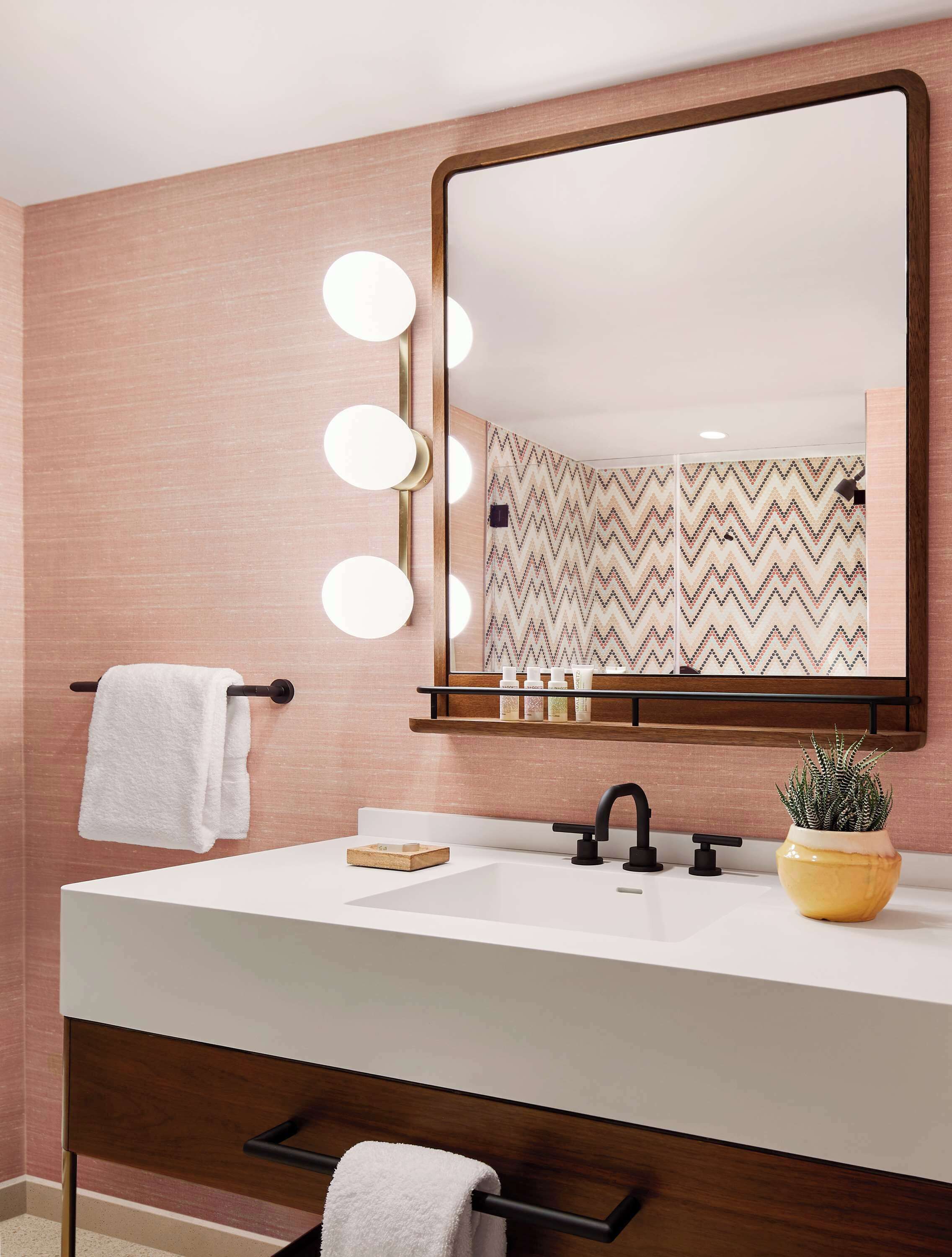 A bathroom with a vanity, a mirror with lights, a towel rack, a shelf with toiletries, and a potted plant.