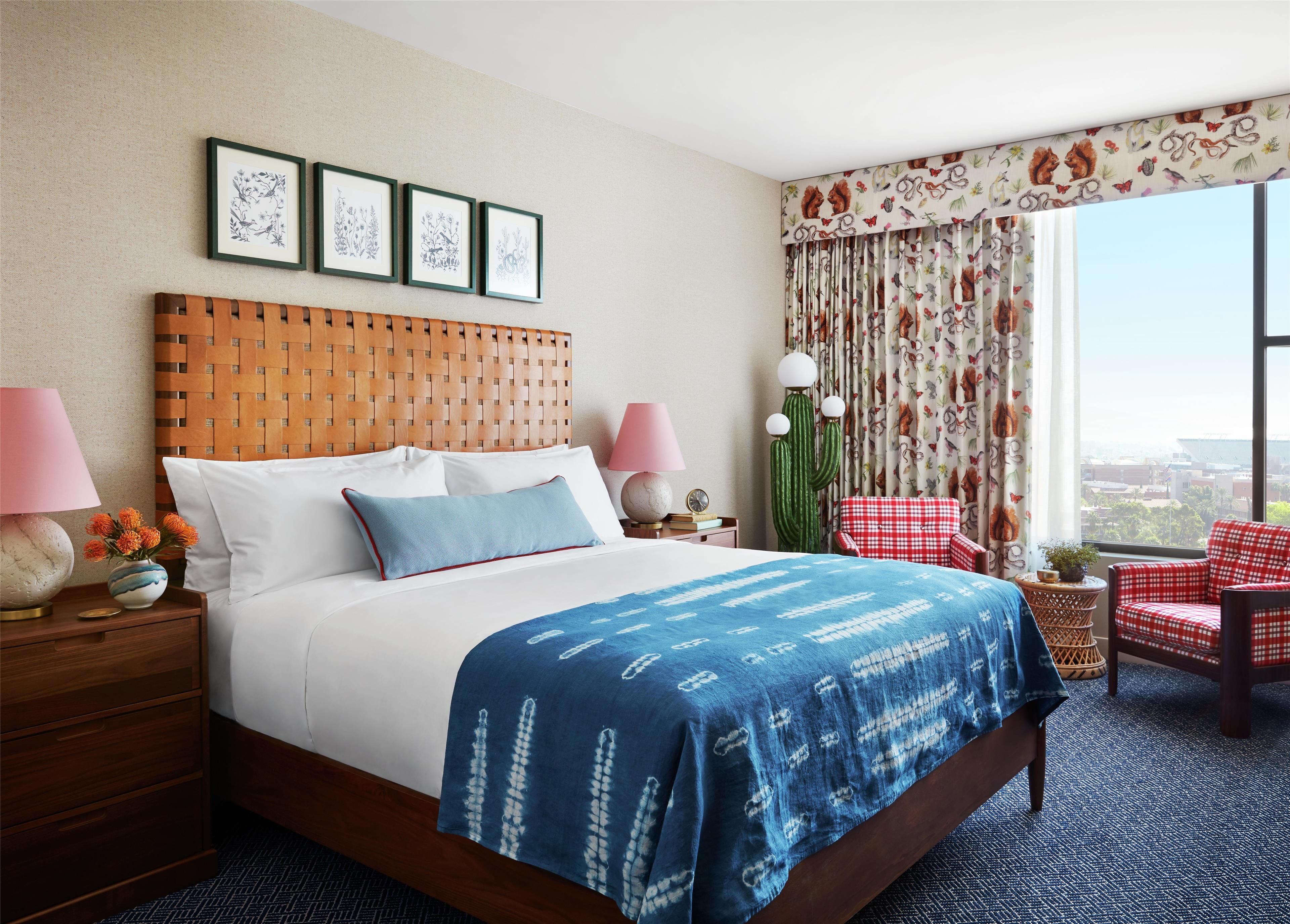 A hotel room with a large bed, a wooden headboard, framed artwork on the wall, a window with curtains, a red checkered chair, and a nightstand with a lamp and decorative items.