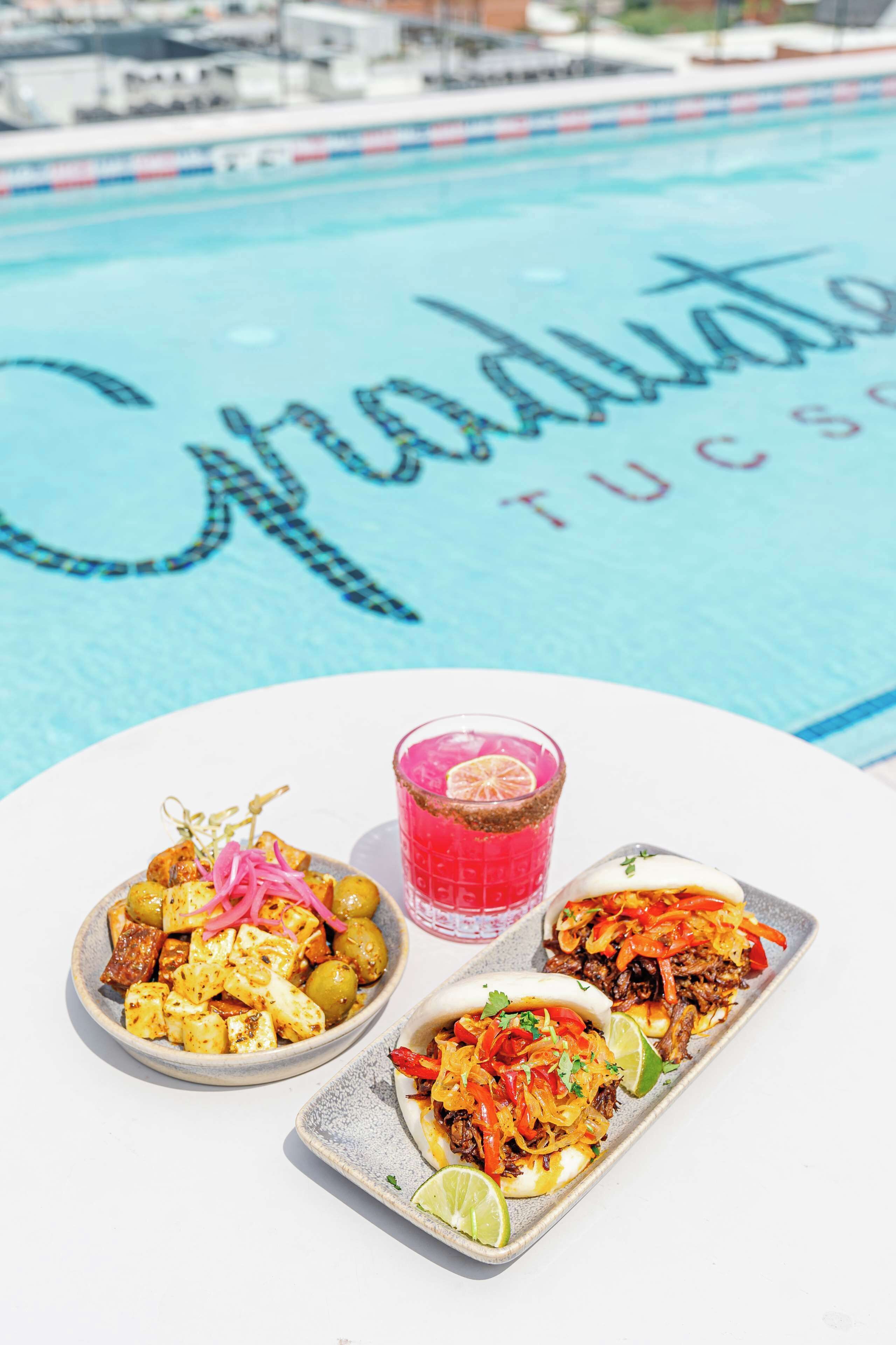 A table with a meal consisting of grilled tofu, a pink cocktail, and a dish with shredded meat and vegetables, set against a pool with a 'Graduate' sign.