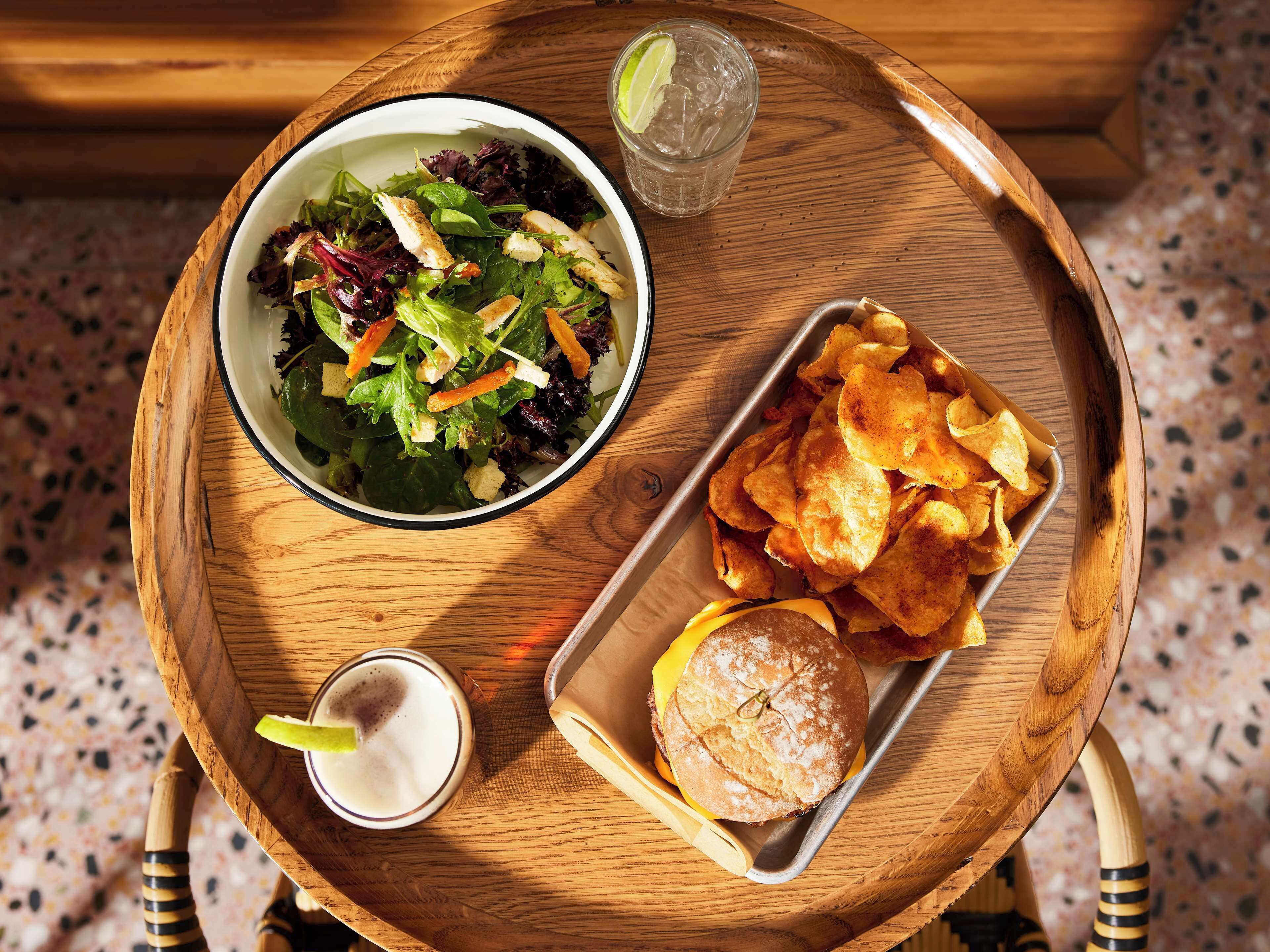 A wooden tray with a salad, chips, a sandwich, and a drink.
