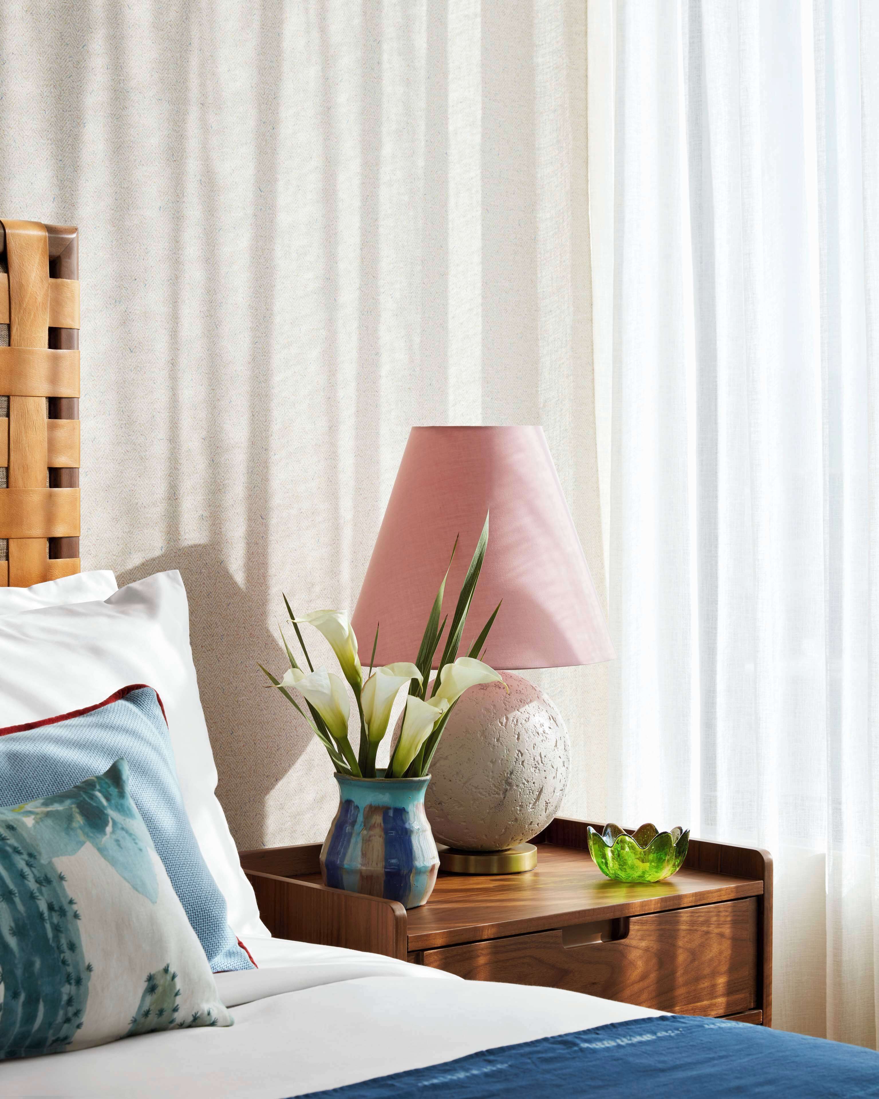A neatly made bed with white and blue bedding, a wooden headboard, a bedside table with a pink lamp, a vase with white flowers, and a decorative stone sphere.