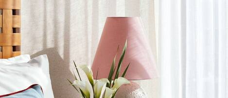 A neatly made bed with white and blue bedding, a wooden headboard, a bedside table with a pink lamp, a vase with white flowers, and a decorative stone sphere.