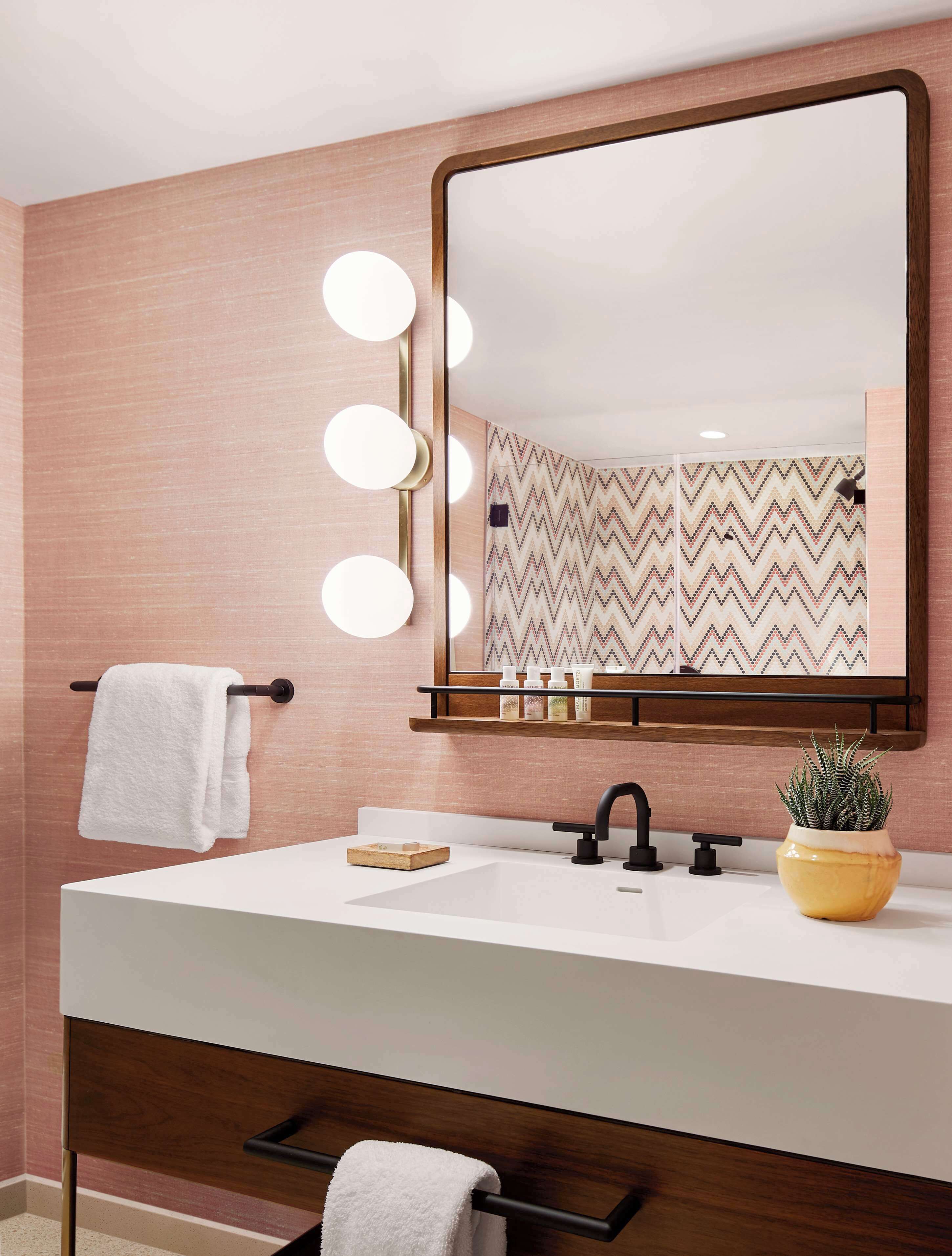 Un baño con tocador, un espejo con luces, toallero, lavabo y planta en maceta.