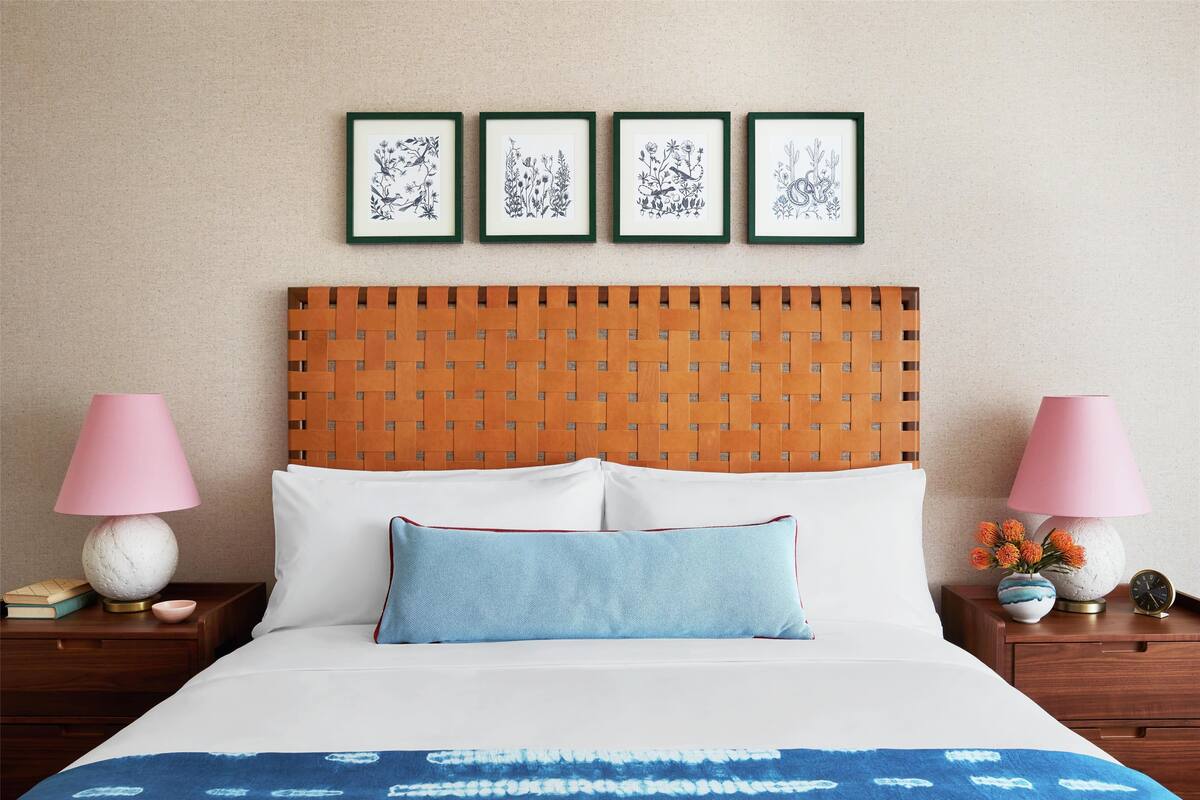 A neatly made bed with a blue patterned bedspread, two pillows, and a wooden headboard. There are framed botanical prints on the wall above the bed.
